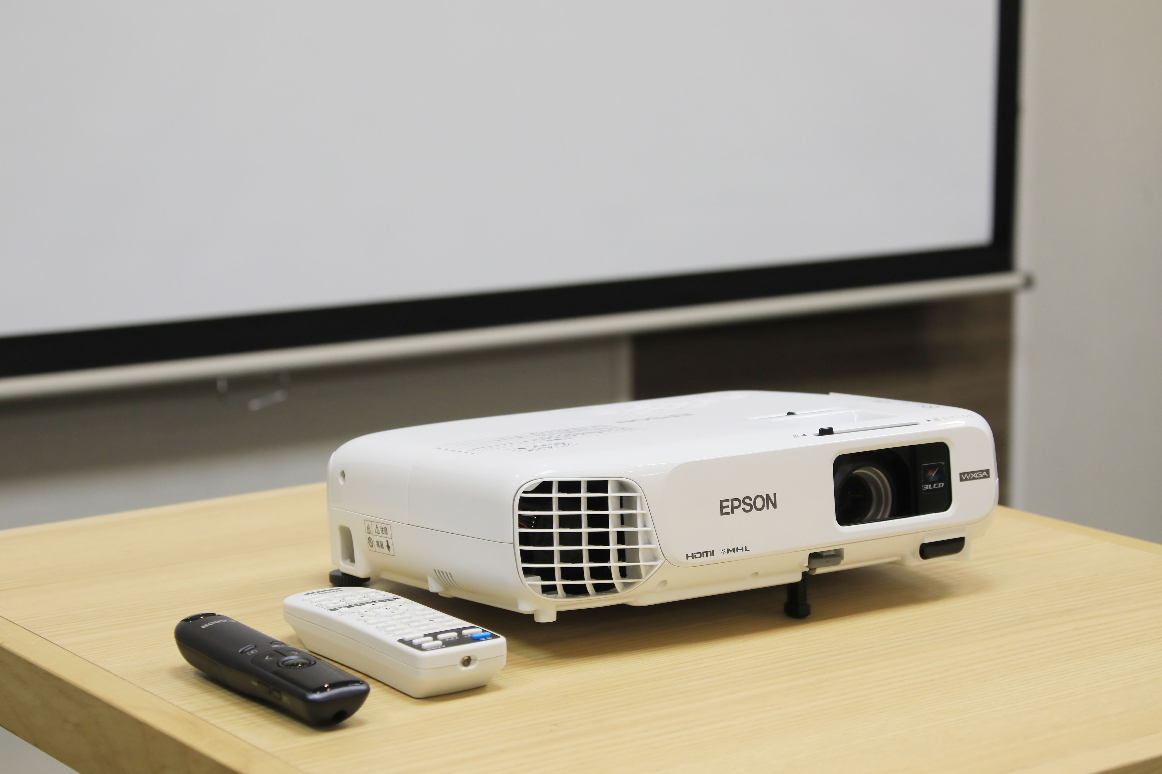 A white Epson projector on a wooden table with a remote control beside it.