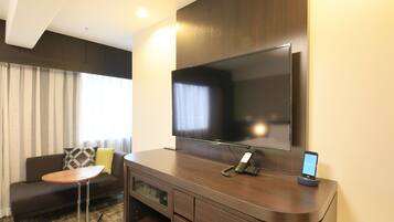 A modern hotel room with a large flat-screen TV mounted on the wall, a sofa with a small round table, and a built-in TV stand with a phone and charging station.
