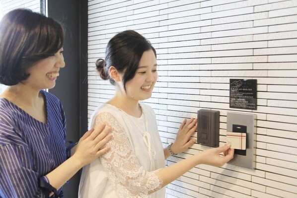 Two individuals interacting with a wall-mounted device, possibly a keycard reader, in a room with a white brick wall.