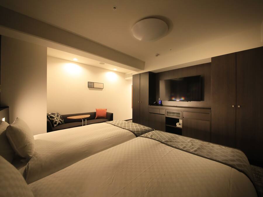 Quarto de hotel moderno com duas camas, uma televisão de tela plana, uma escrivaninha e uma área de estar com sofá e cadeiras.