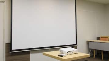 Un salón de conferencias con una pantalla de proyección grande, un proyector y un control remoto sobre una estantería de madera.