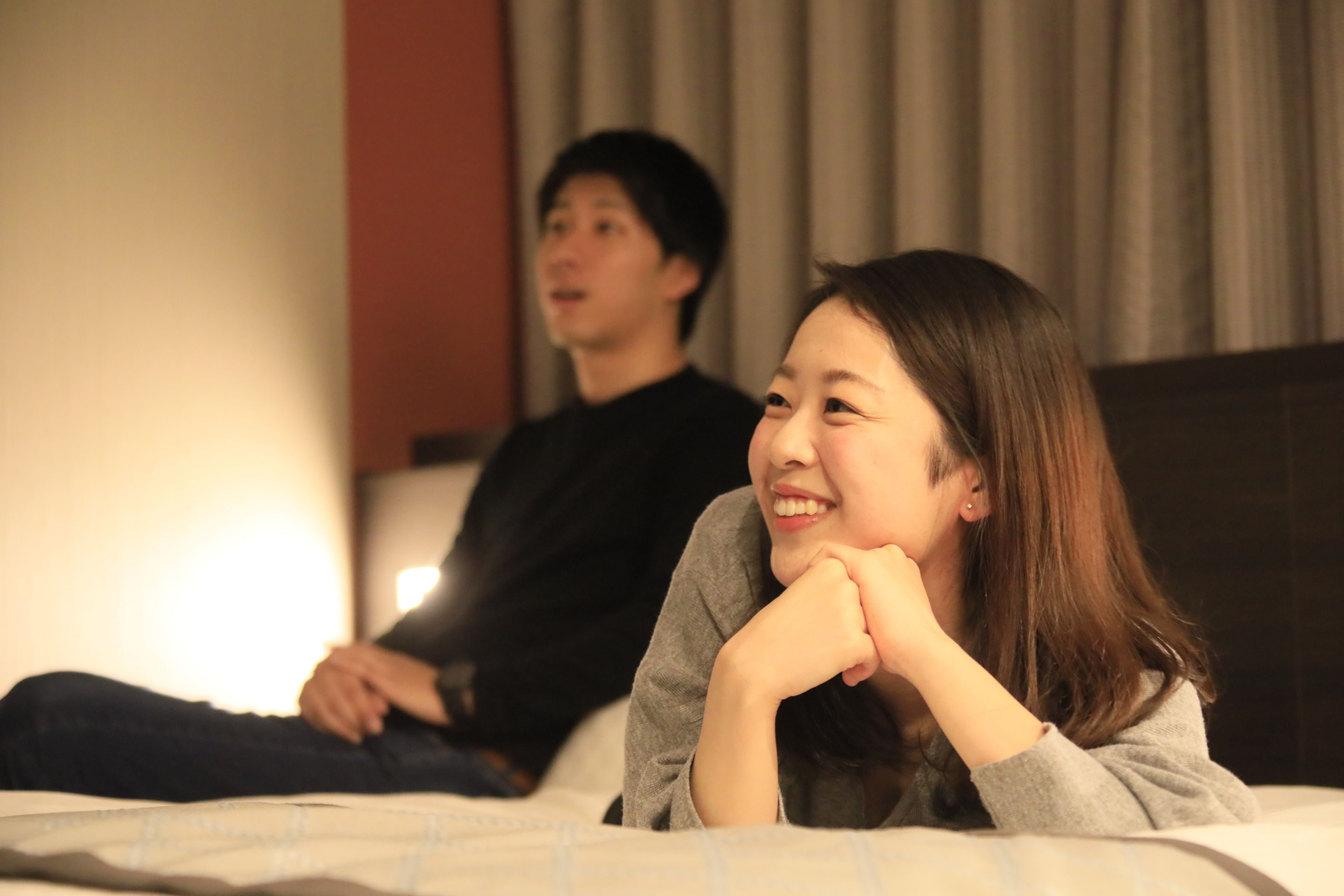 A room with a bed, a person sitting on the bed, and another person standing in the background.