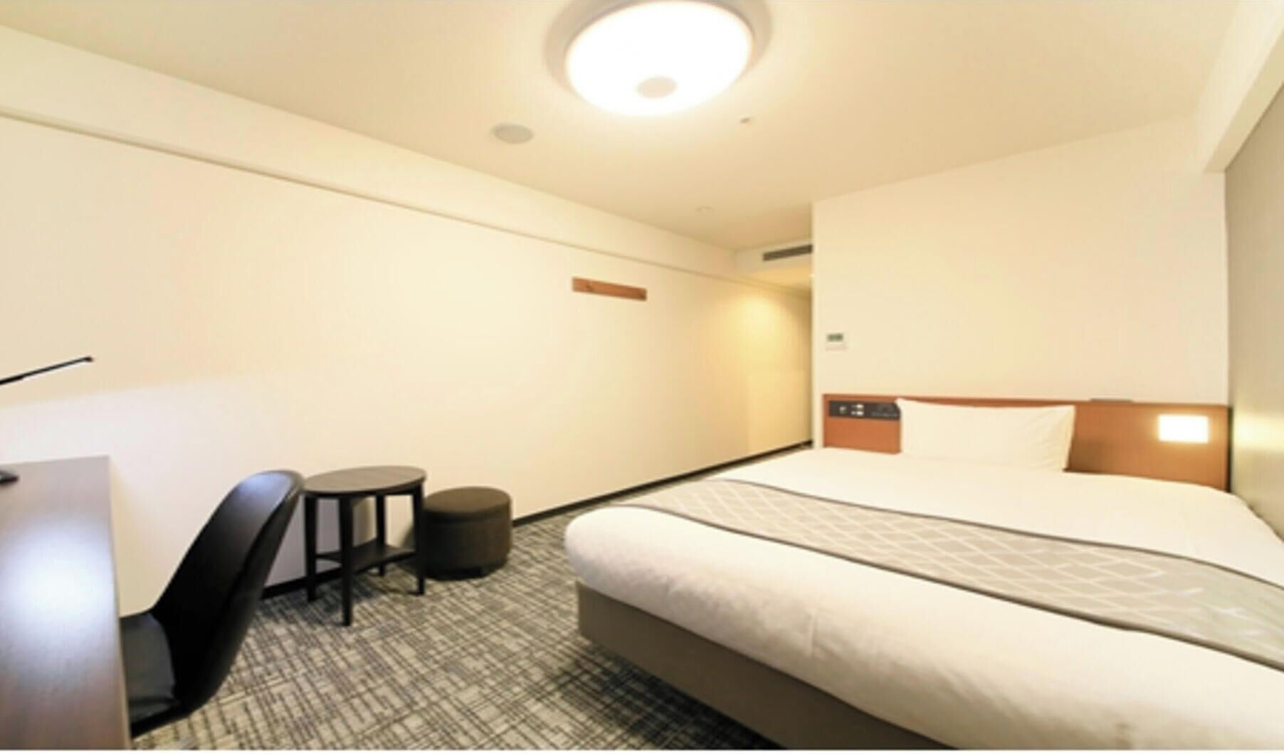 A hotel room with a bed, a desk, a chair, and a small stool.