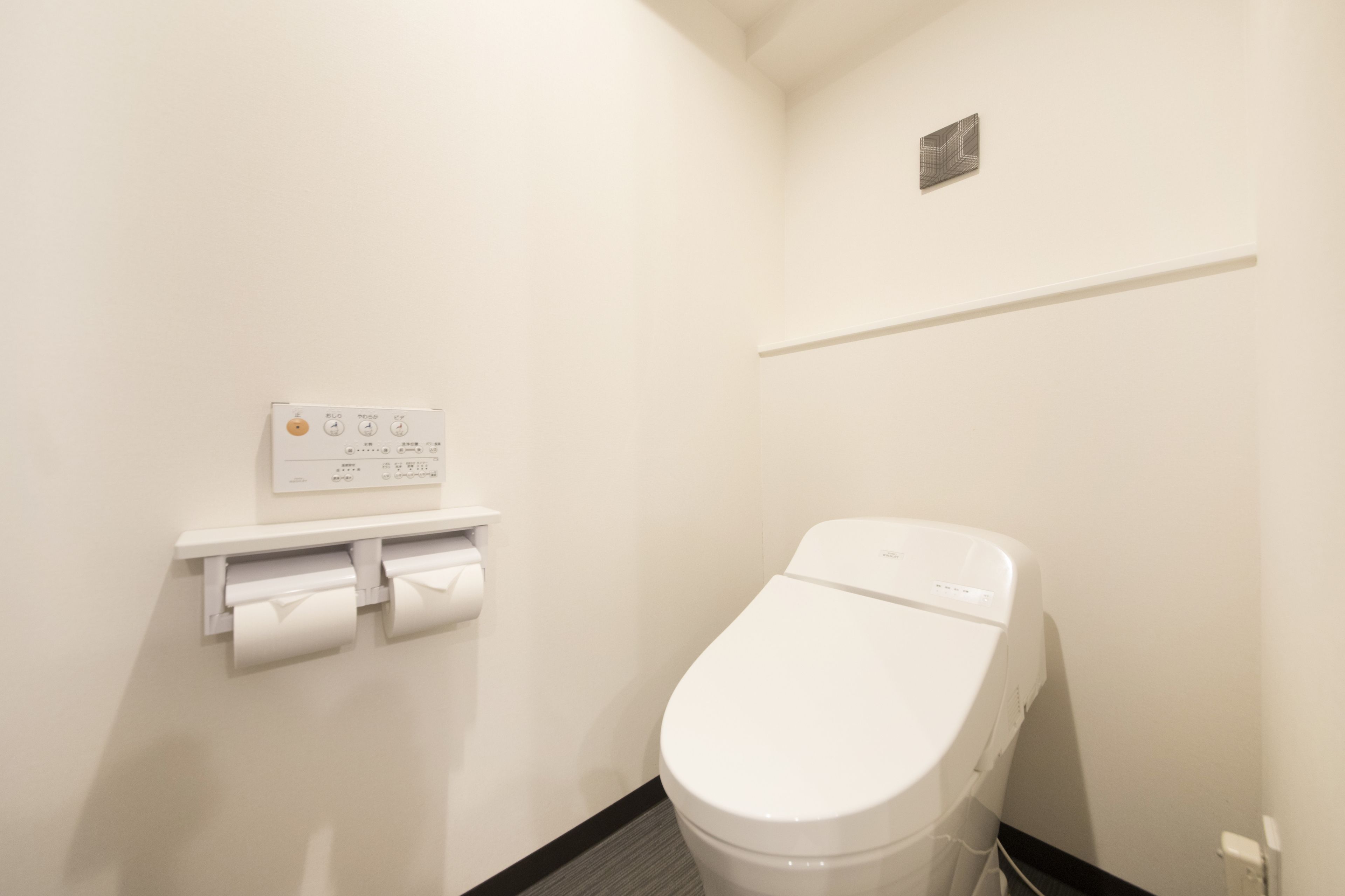 A small, clean bathroom with a toilet, a toilet paper holder, and a wall-mounted flush control panel.