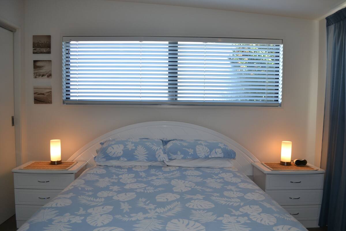 A bedroom with a bed, two bedside tables, a window with blinds, and wall-mounted pictures.