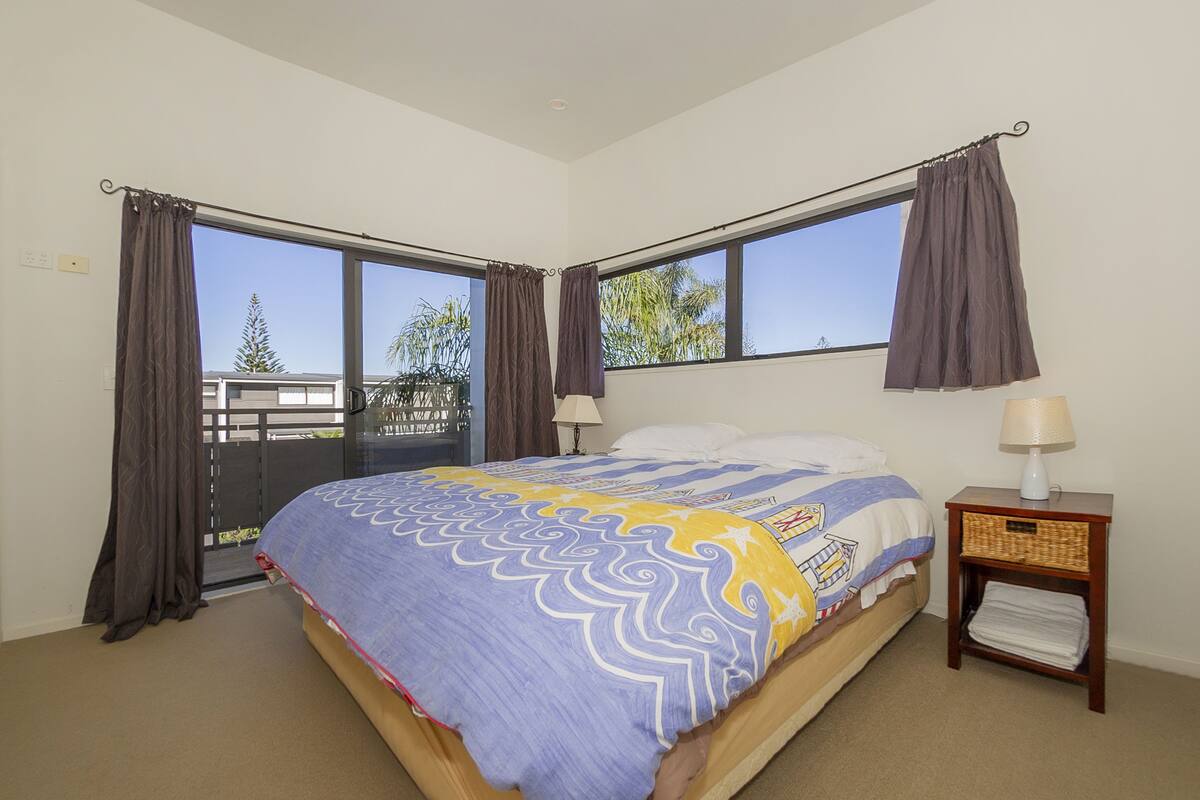 A bedroom with a bed, two windows, a nightstand, and a view of the outside.