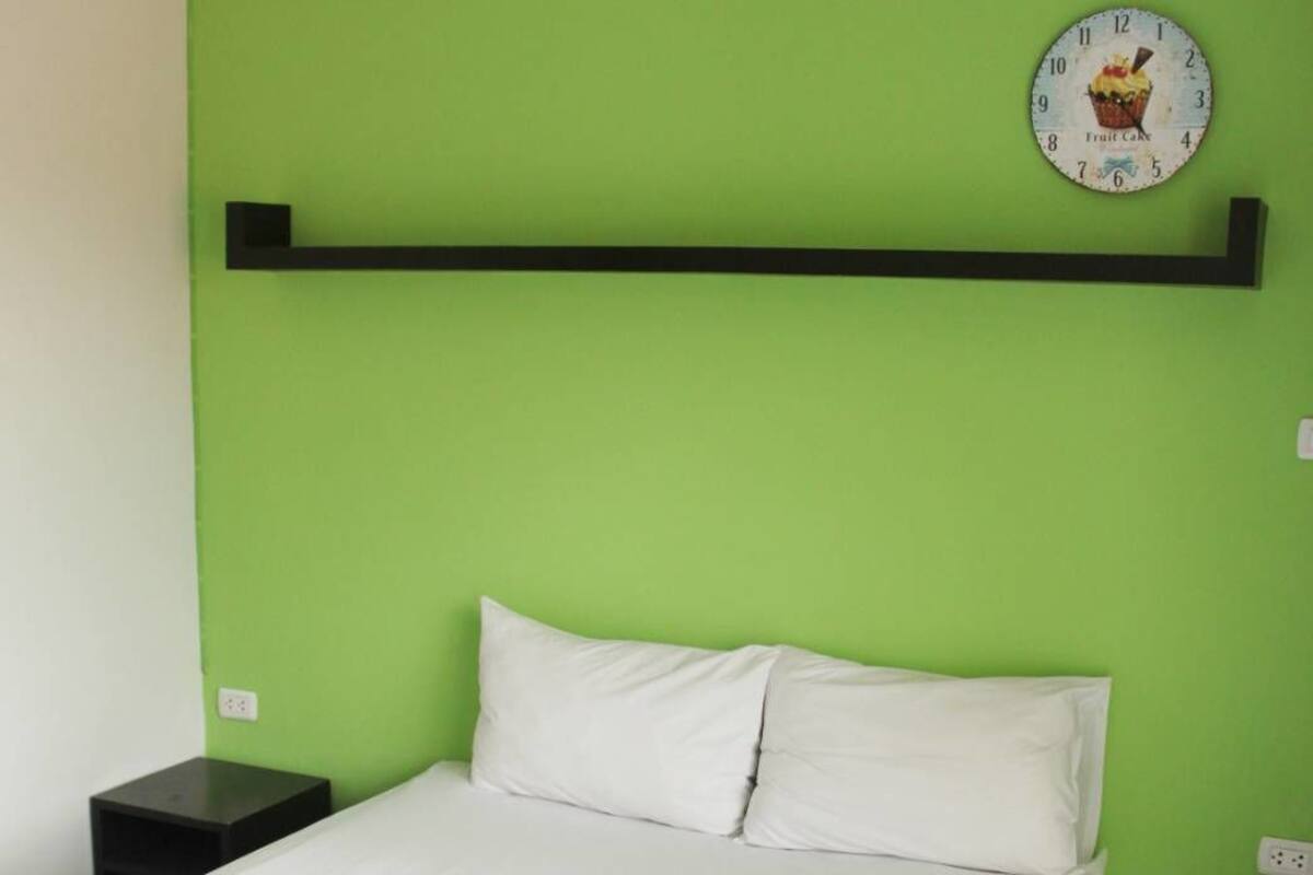Habitación de hotel con pared verde, cama con ropa de cama blanca, mesita de noche negra y un reloj de pared con diseño de cupcake.