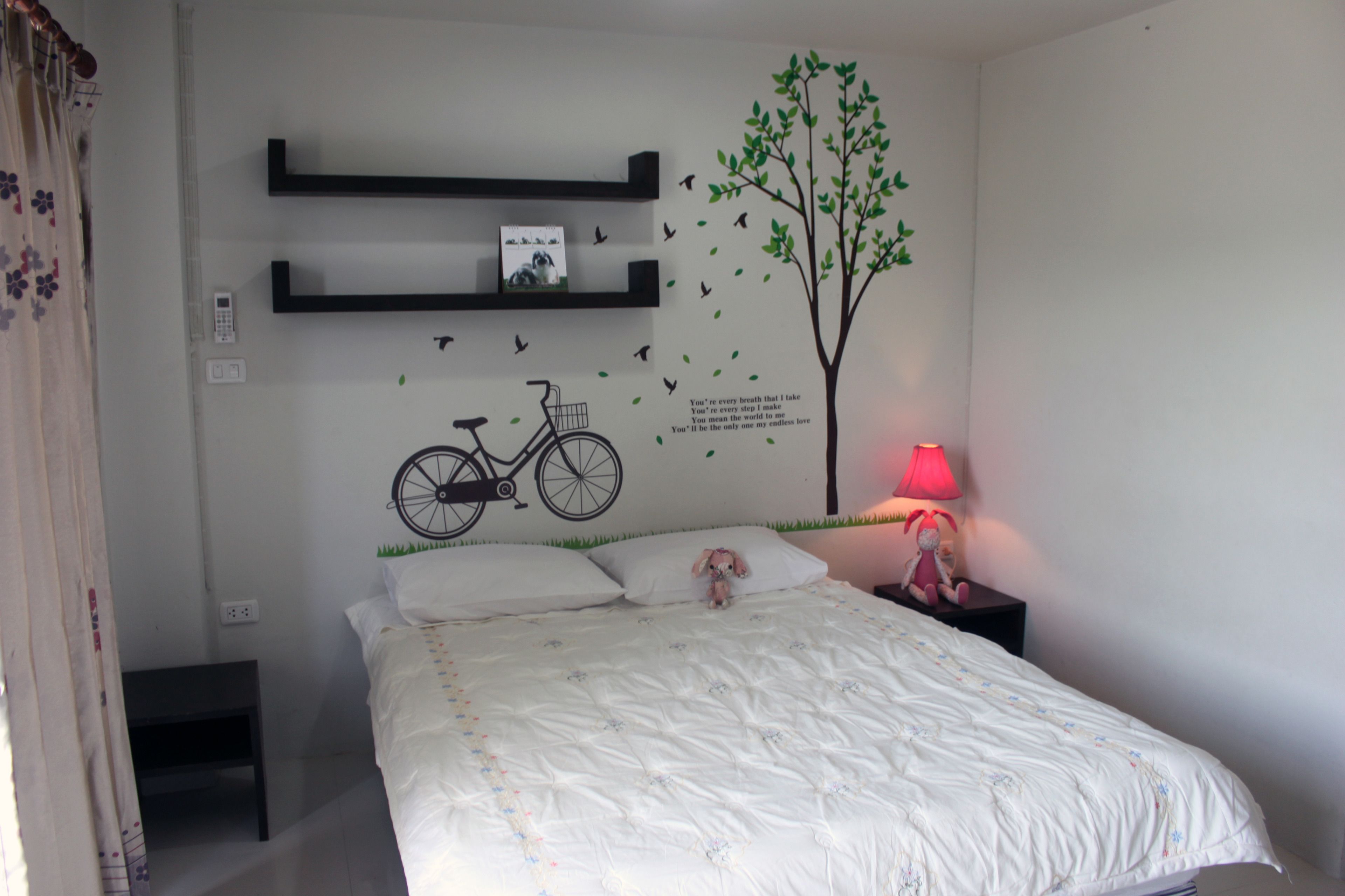 Un dormitorio con cama, una mesita de noche, una lámpara, un adhesivo mural con un dibujo de una bicicleta y un árbol, y una estantería con un cuadro y un teléfono.
