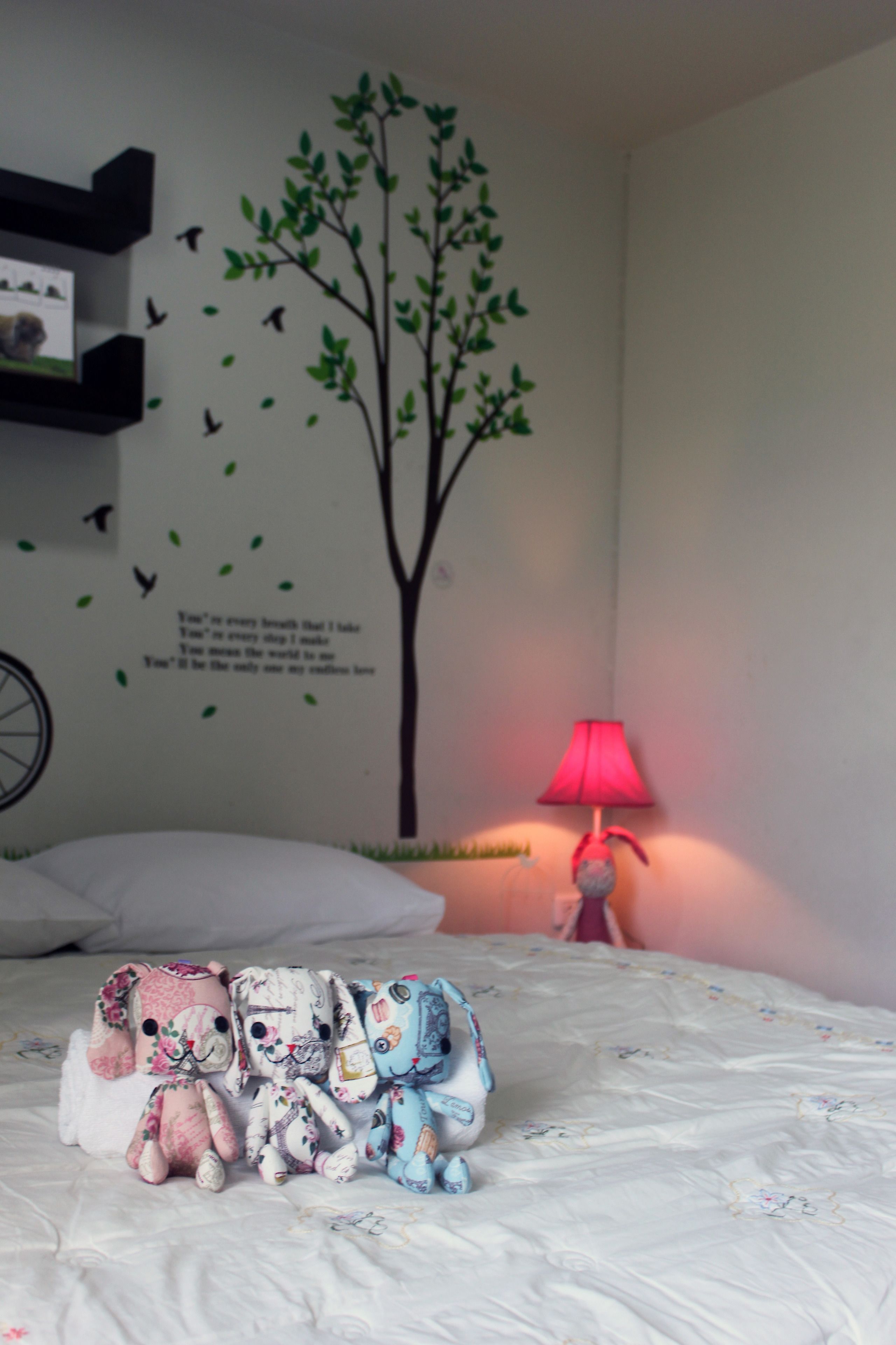 Une chambre à coucher avec un lit, un motif décoratif représentant un arbre et des oiseaux sur un mur, une lampe et trois peluches sur le lit.