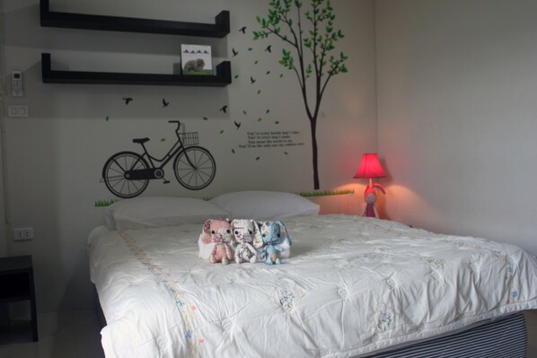 A neatly made bed with a white comforter and a colorful blanket, a wall decal of a bicycle and tree, a red lamp on a bedside table, and a small shelf above the bed.