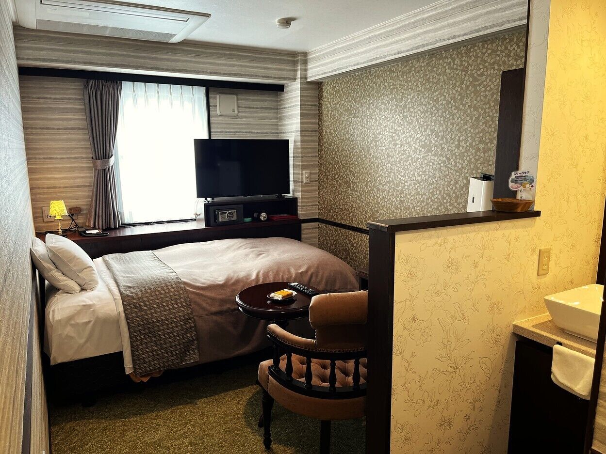 A hotel room with a bed, a TV, a small table with a chair, a bathroom with a sink, and a mirror.