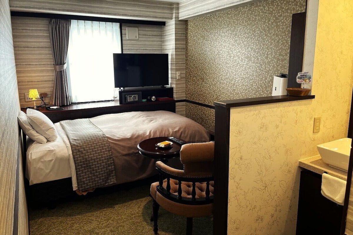 A hotel room with a bed, a TV, a small table with a chair, a bathroom with a sink, and a mirror.