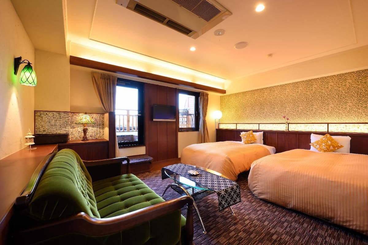 A hotel room with two beds, a green armchair, a small table, a TV, and a window with curtains.