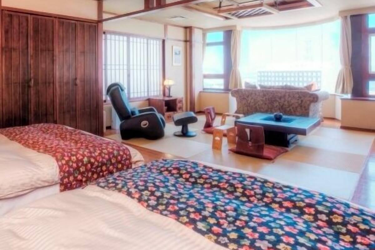 A hotel room with a large bed, a sofa, a massage chair, and a view of the city.