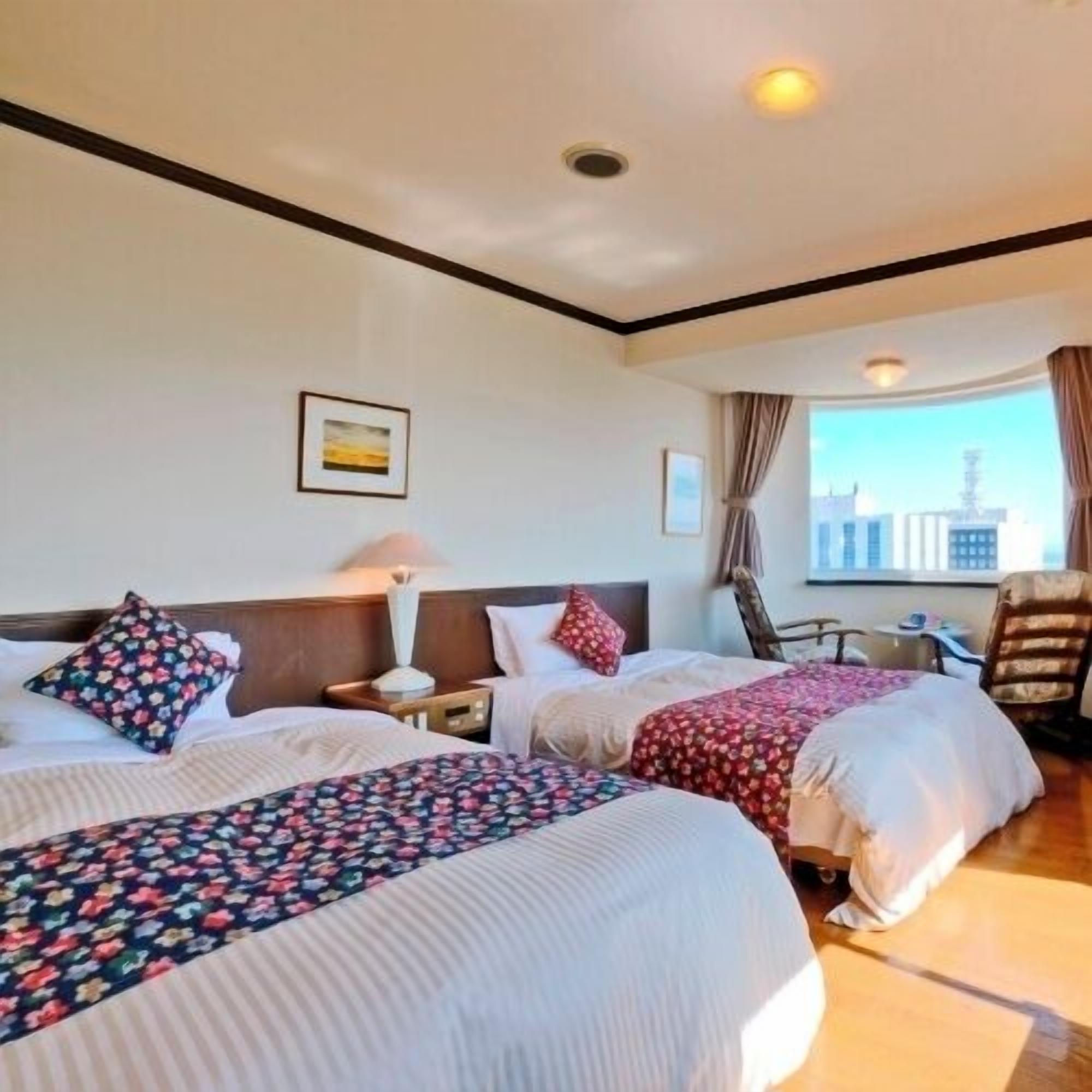 A hotel room with two beds, a chair, a table, and a window with a city view.