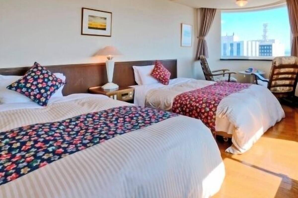 A hotel room with two beds, a chair, a table, and a window with a city view.