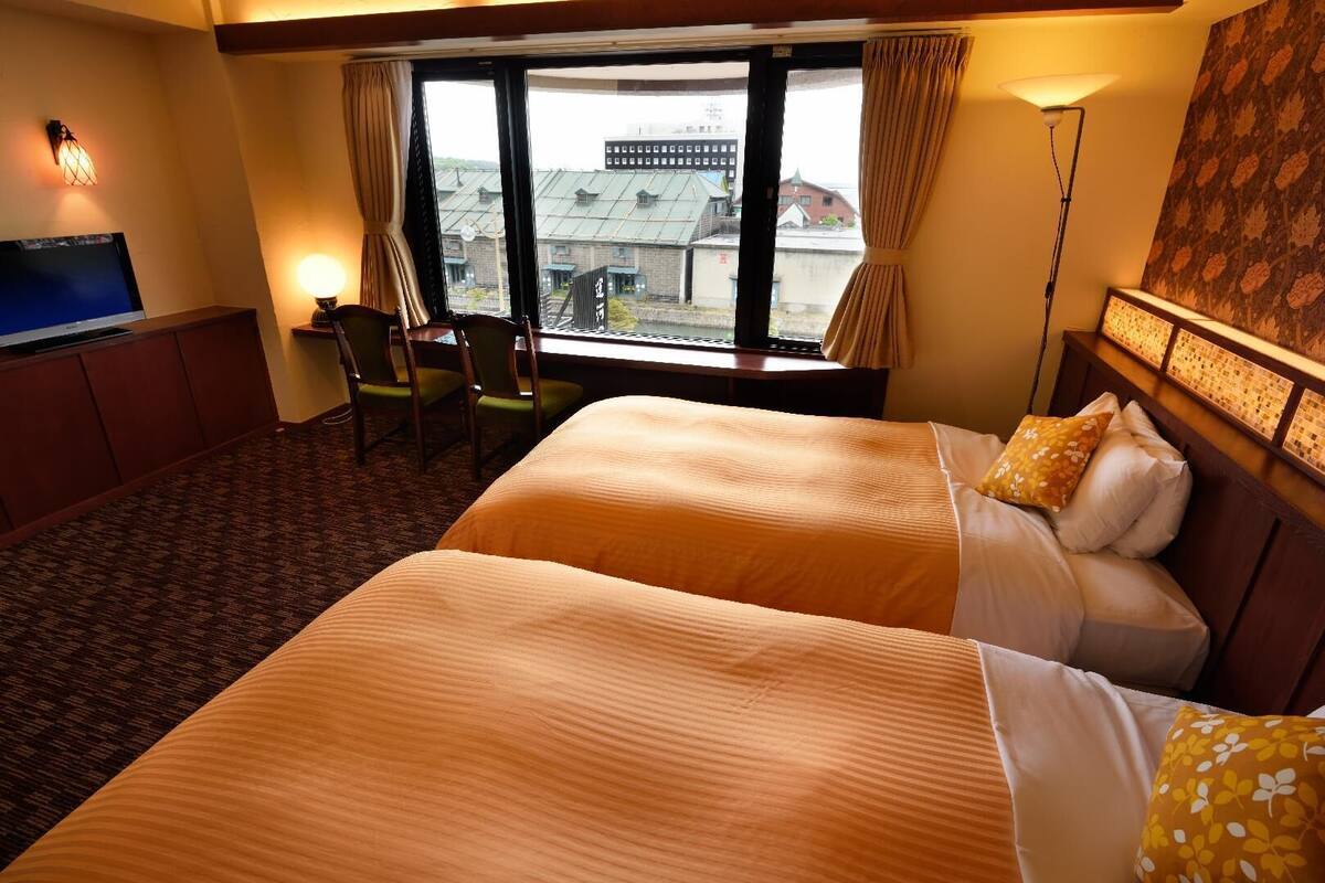 A hotel room with two beds, a desk, a chair, a TV, and a large window with a view of buildings.