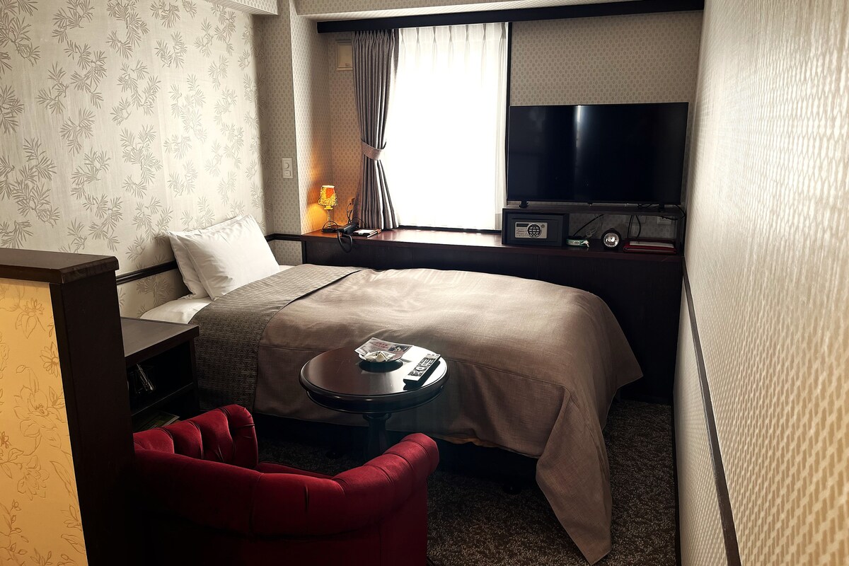 A hotel room with a bed, a TV, a desk with a lamp, a red chair, and a small table.