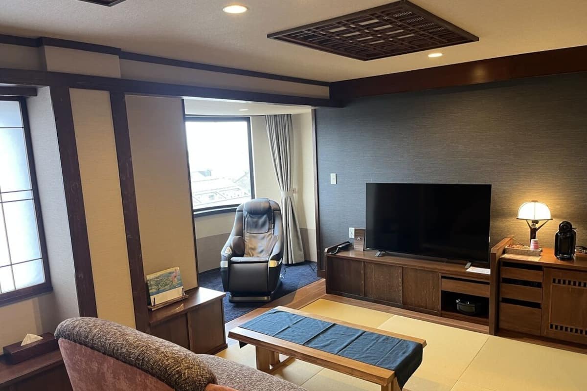 A hotel room with a sofa, a massage chair, a TV, a small table, and a window with curtains.