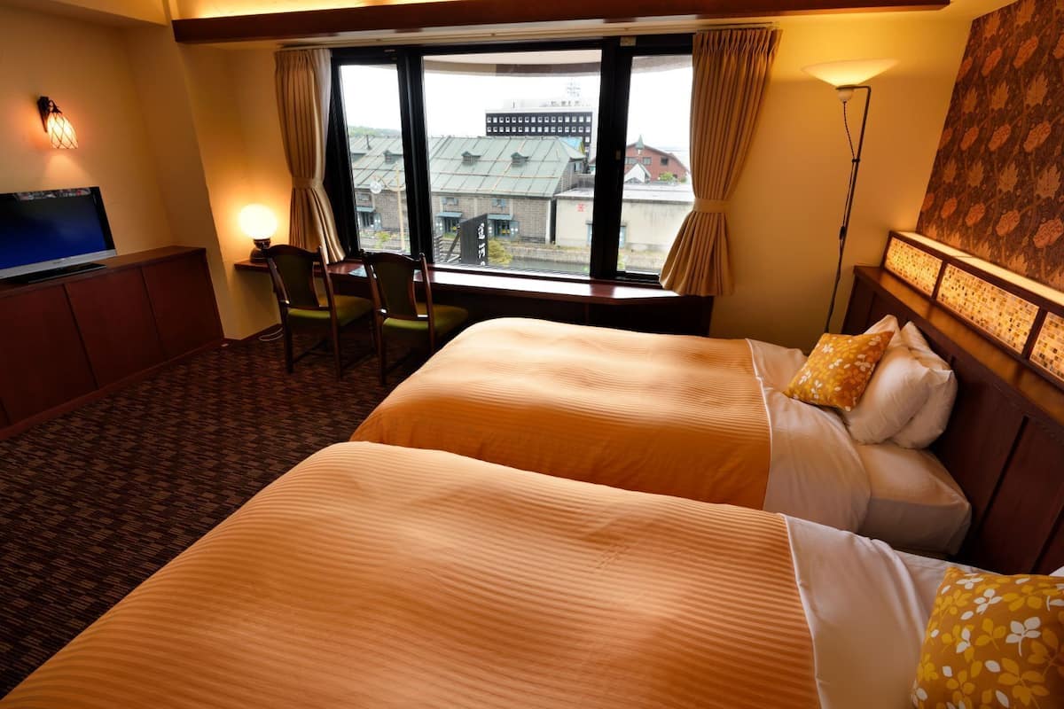 A hotel room with two beds, a desk, a chair, a TV, and a view of buildings through the window.