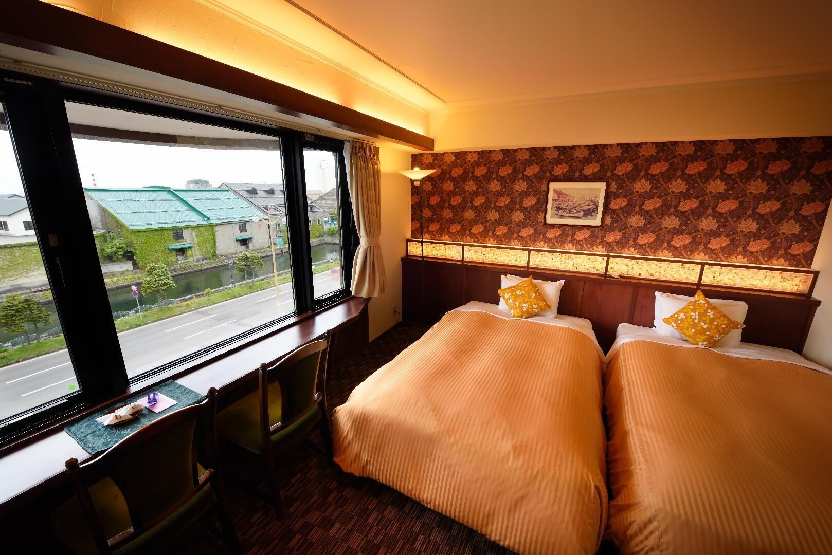 A hotel room with two beds, a desk, and a window with a view of a street and buildings.