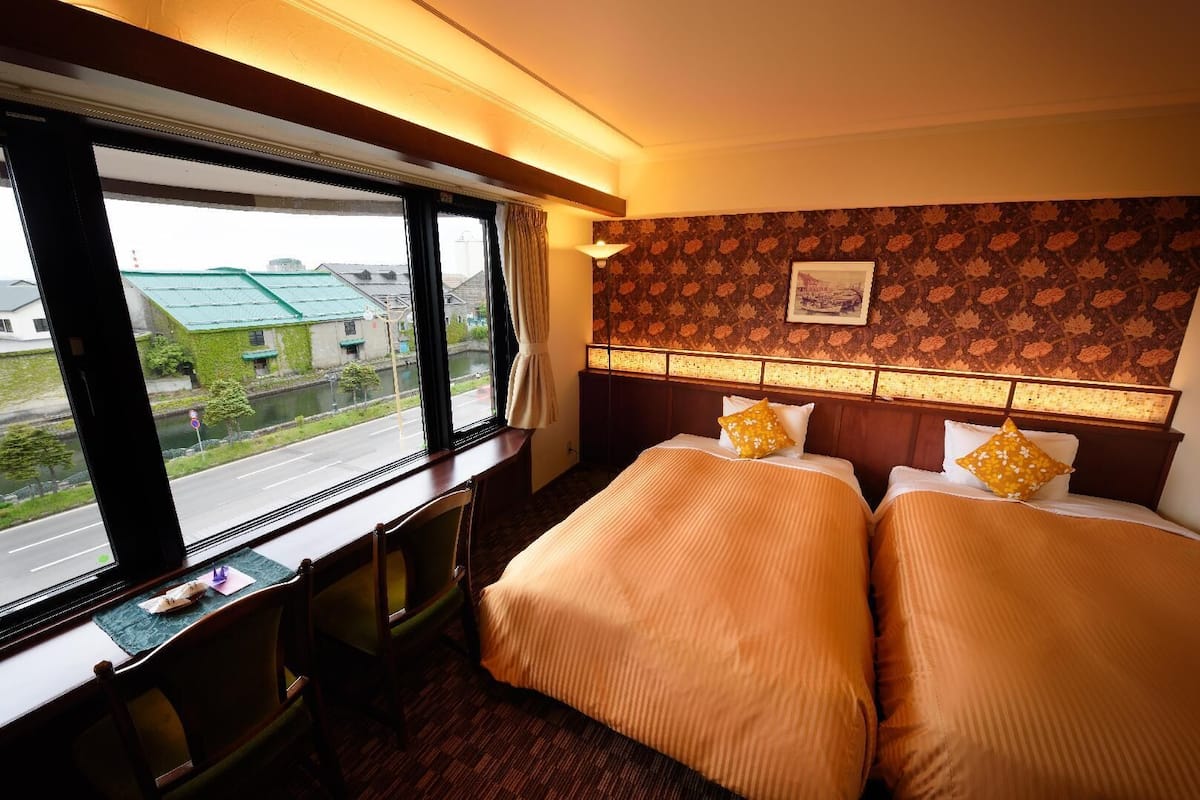 A hotel room with two beds, a desk, and a window with a view of a street and buildings.