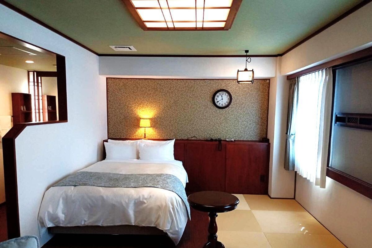 A hotel room with a bed, bedside table, wall clock, and a window with curtains.