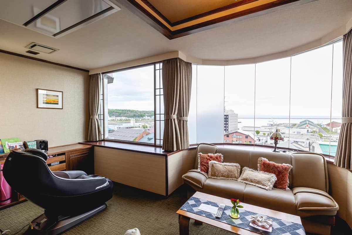 A hotel room with a leather sofa, a black chair, a desk with a lamp, and a large window offering a city view.
