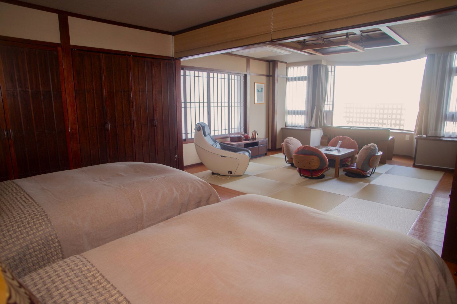 A hotel room with two beds, a massage chair, a small dining area, and large windows.