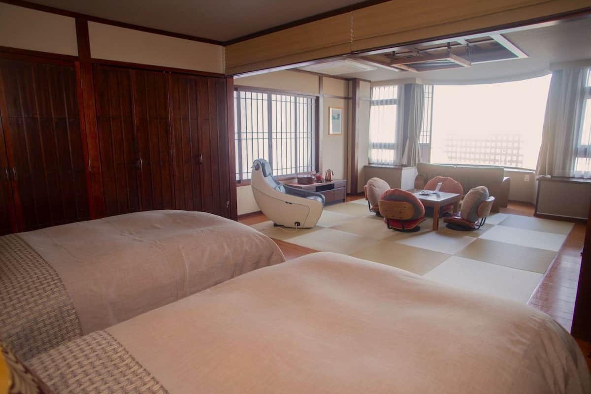 A hotel room with two beds, a massage chair, a small dining area, and large windows.