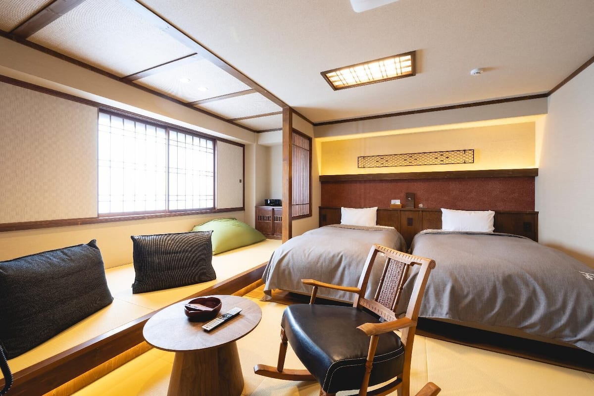 A hotel room with a traditional Japanese interior, featuring tatami flooring, a low wooden bed, a futon, a wooden chair, a small table, and a window with a view.