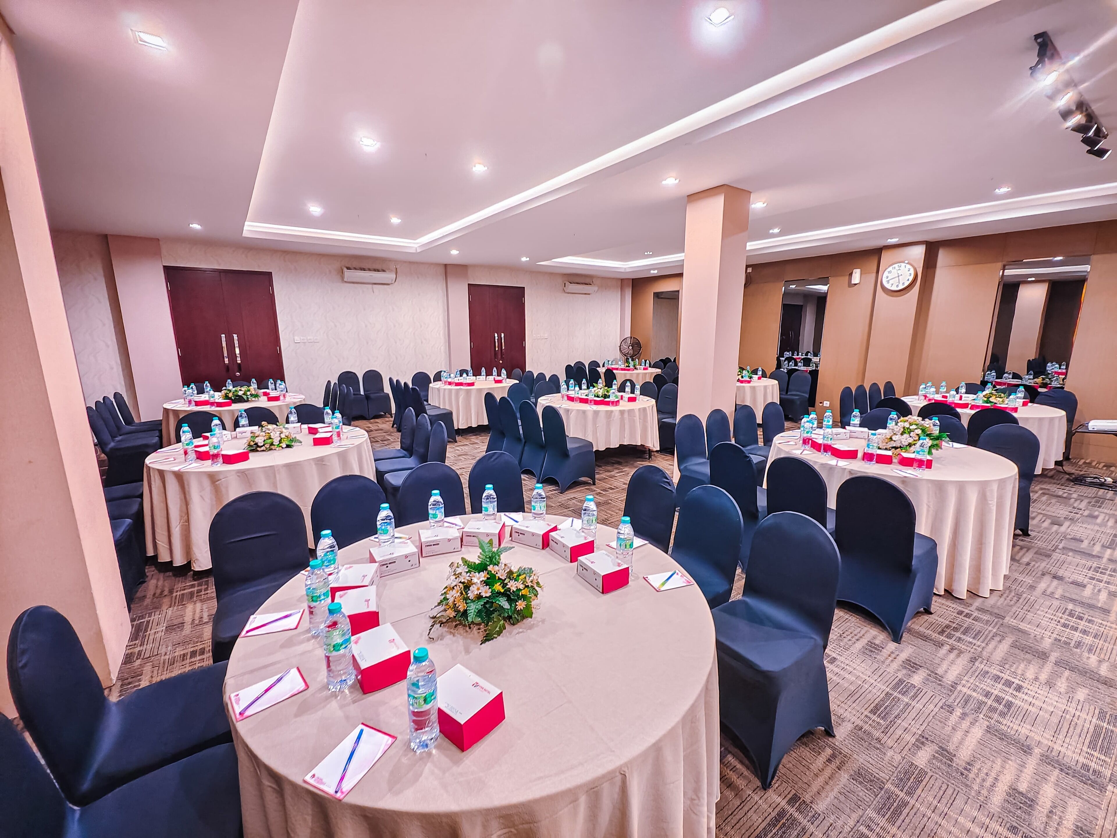 Un salón de conferencias con varias mesas redondas, cada una con botellas de agua, tarjetas de identificación y centros de mesa florales.