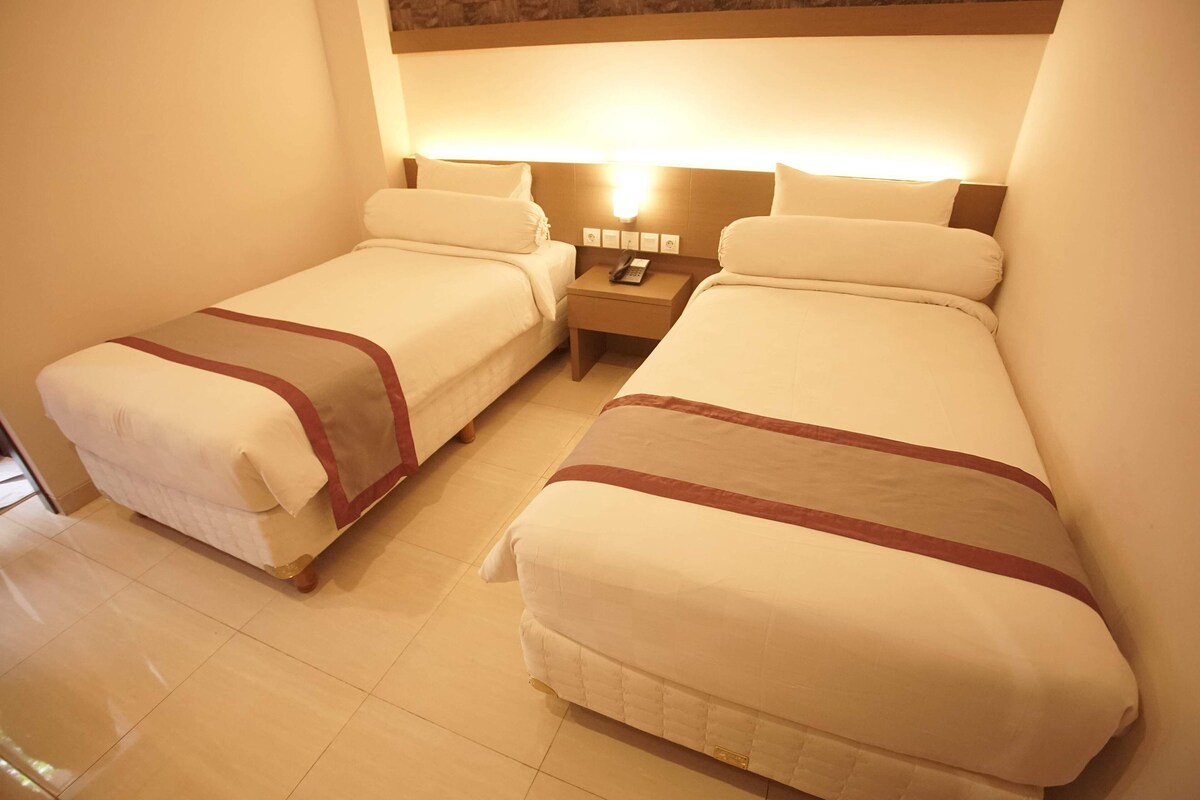 A hotel room with two beds, a nightstand with a phone, and a wall-mounted light.