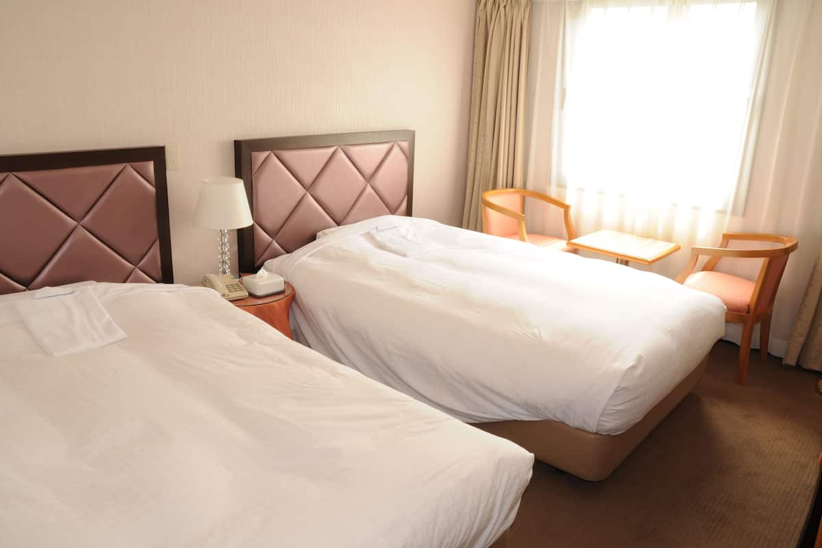 Habitació d'hotel amb dos llits, una taula de nit amb una làmpada, una cadira i una finestra amb cortines.