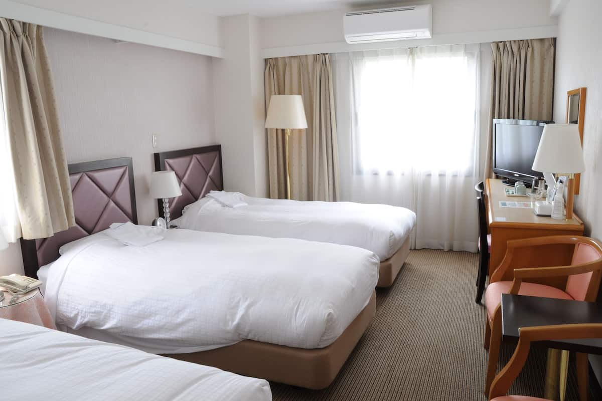 Habitació d'hotel amb dos llits, un escriptori, una cadira, un televisor i una finestra amb cortines.