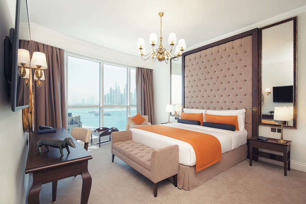 A hotel room with a large bed, a desk, a chair, a small table, a mirror, and a view of the city.