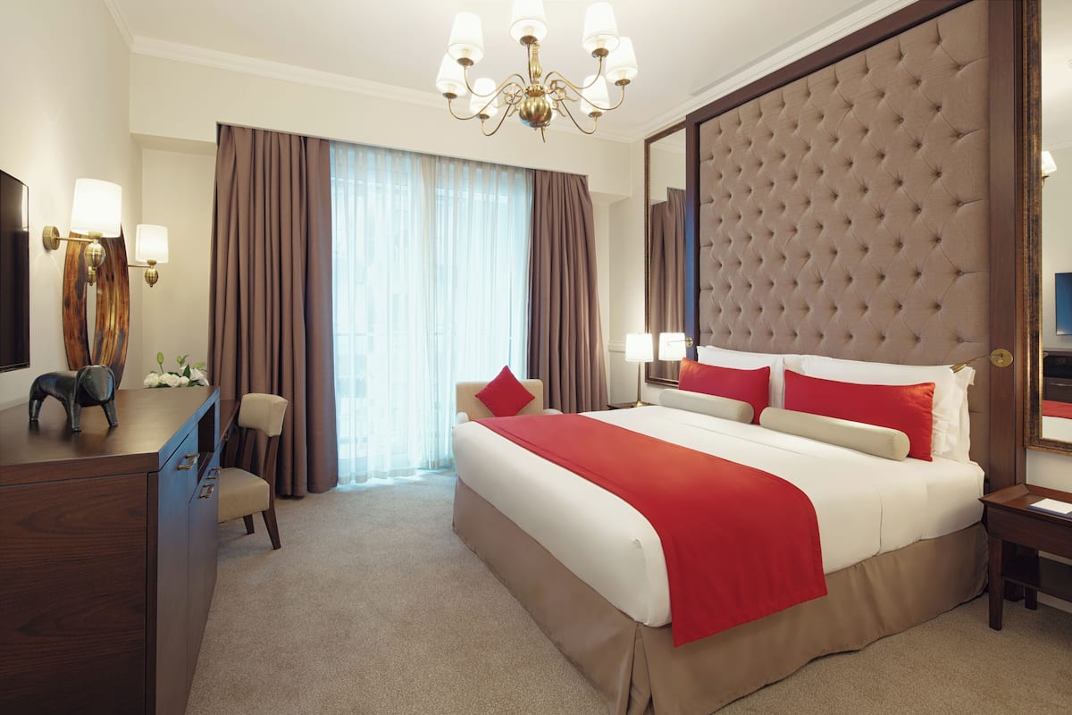 A hotel room with a large bed, a wooden desk, a chair, a desk lamp, a mirror, and a chandelier.