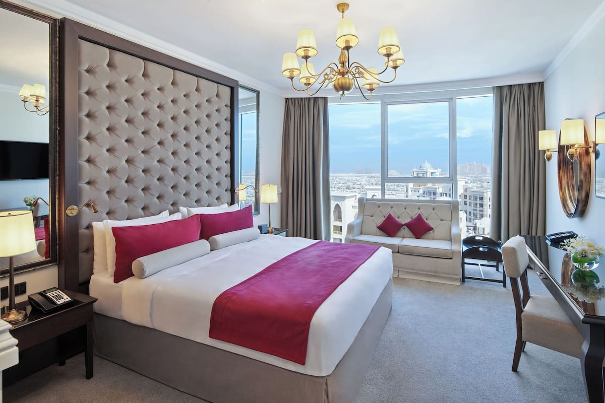 A hotel room with a large bed, a view of the city, a chandelier, and a desk with a lamp.