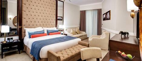 A hotel room with a large bed, a sitting area with a sofa and armchairs, a wooden side table with a lamp, and a small table with fruit.