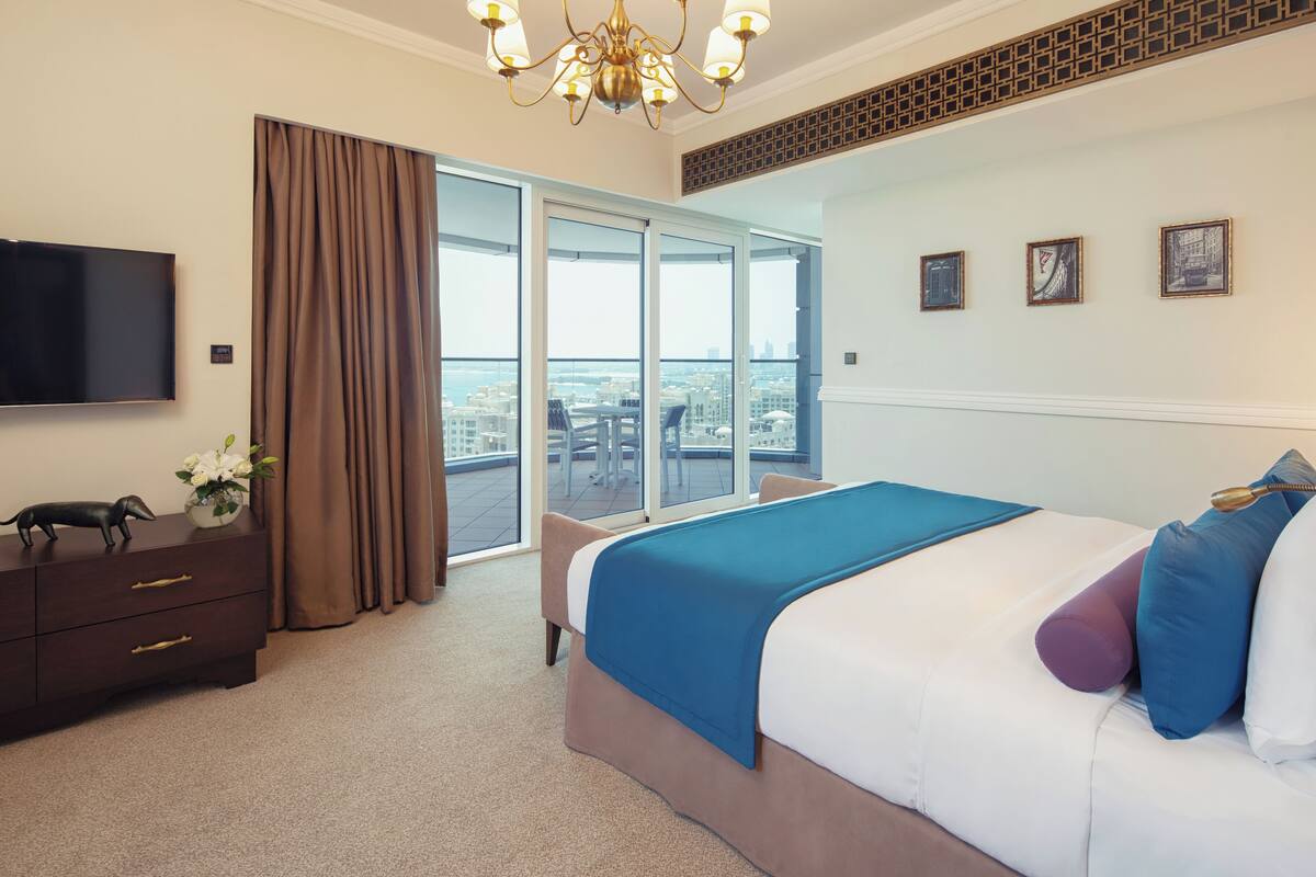 A hotel room with a large bed, a flat-screen TV, a desk with drawers, a chair, a balcony with a view of the city, and a chandelier.