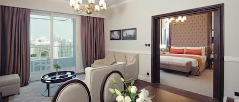 A hotel room with a dining area, a sofa, a round table with a vase of flowers, and a view of the city through large windows.