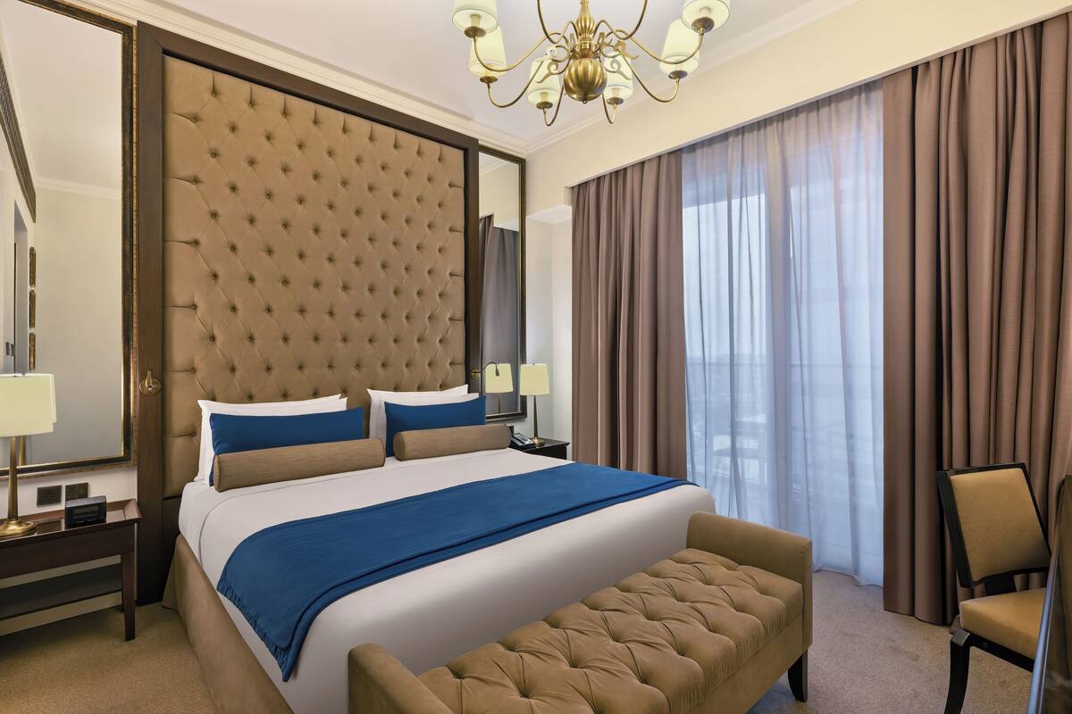 A hotel room with a large bed, a tufted headboard, a chandelier, a sitting area, and a view through sheer curtains.