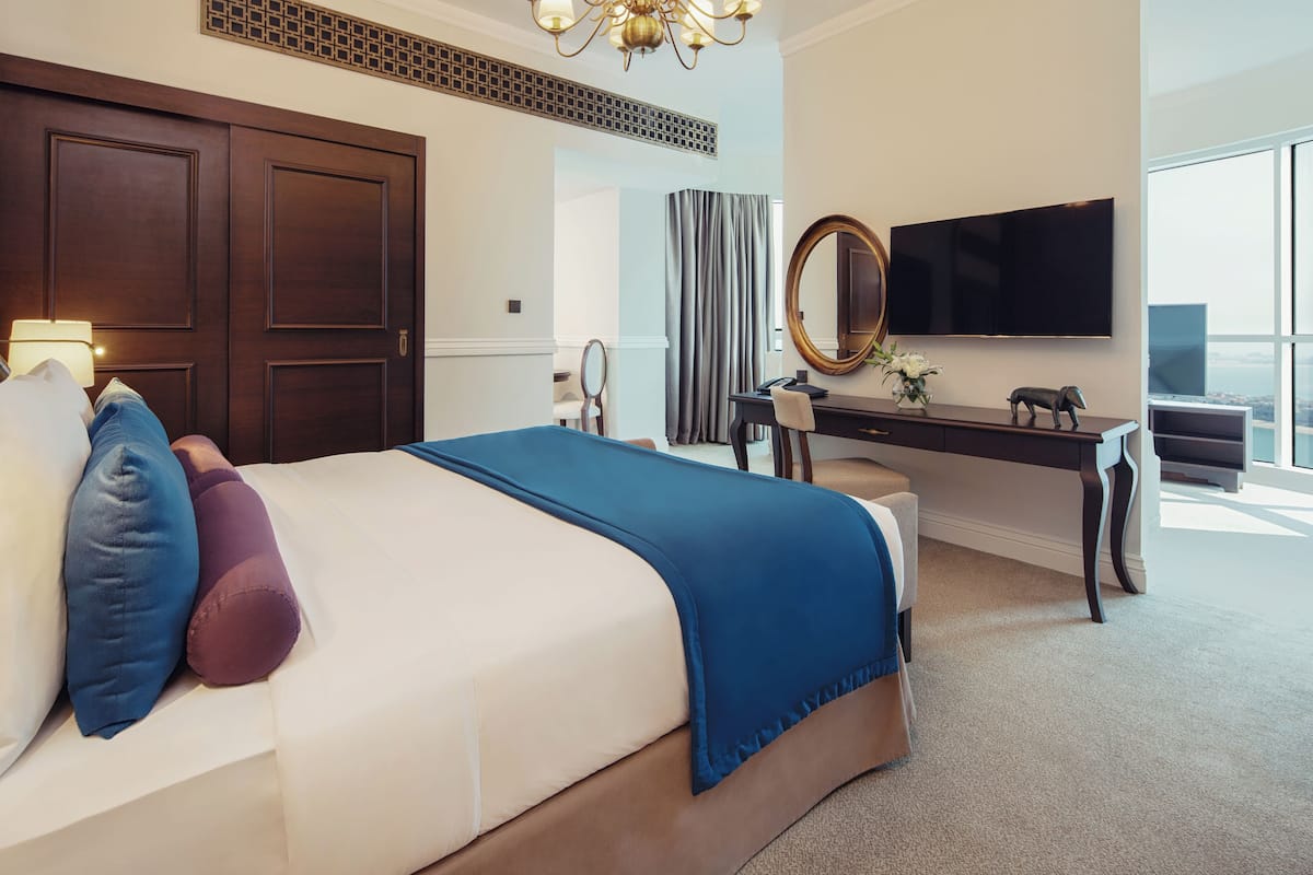 A hotel room with a large bed, a desk with a mirror, a flat-screen TV, and a view of the city.