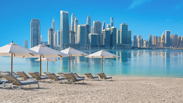 Una playa con sillas reclinables y sombrillas, un cielo despejado y un horizonte urbano al fondo.
