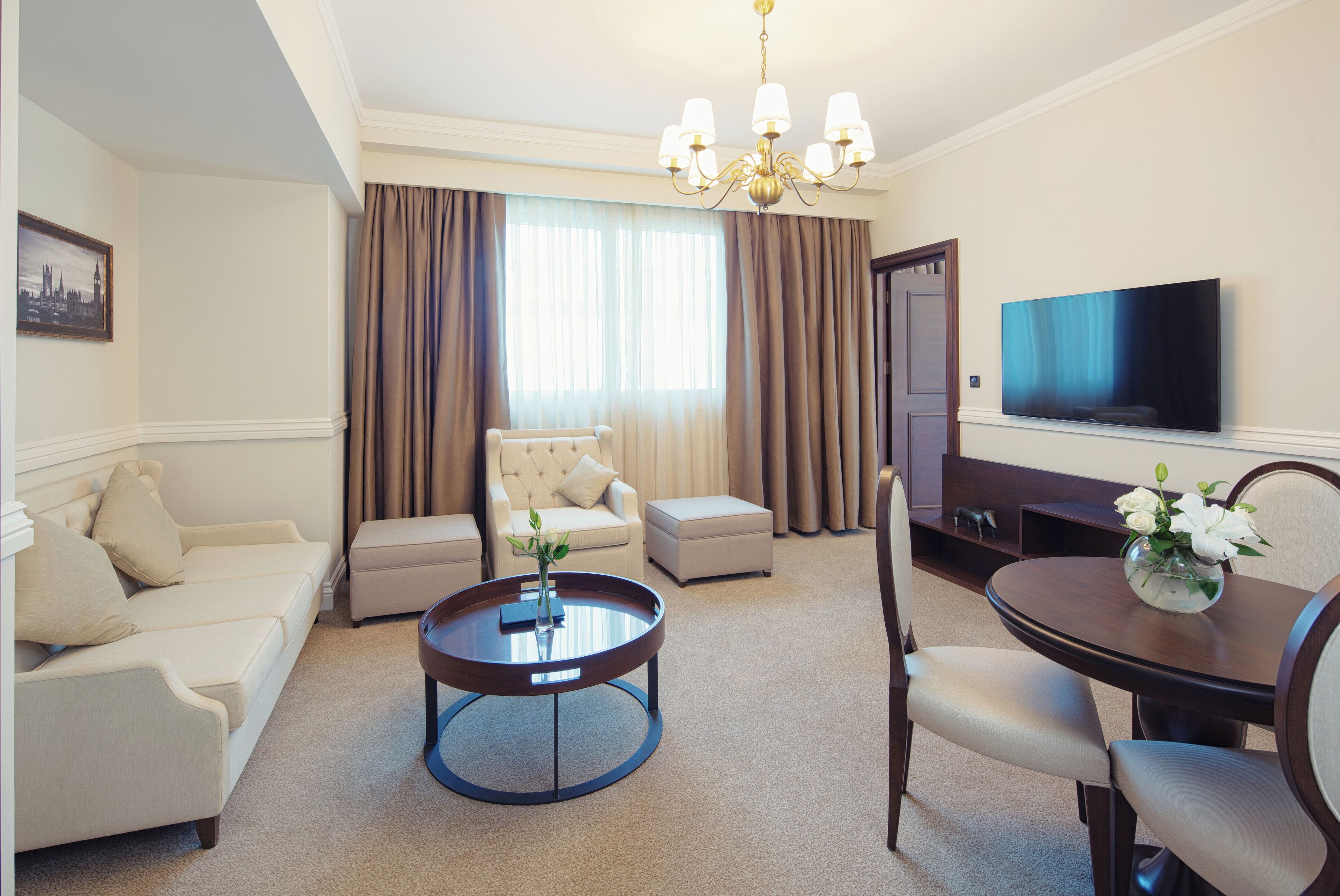 A hotel room with a beige sofa, a round coffee table, a dining table with chairs, a TV, and a large window with curtains.