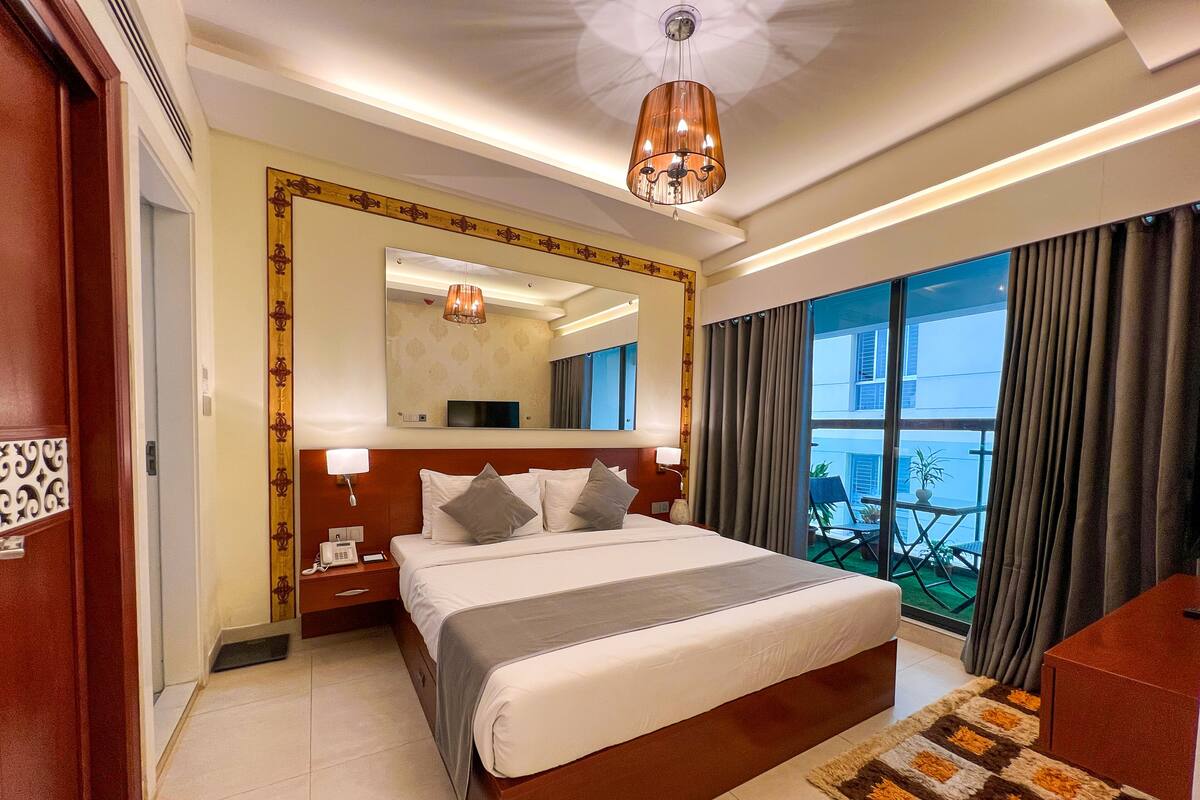 A hotel room with a large bed, a bedside table with a lamp, a mirror, and a view of the outside balcony.
