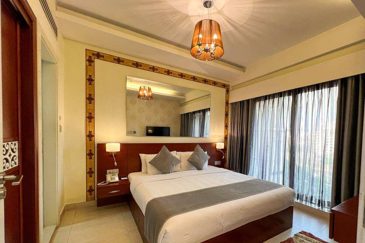 A hotel room with a large bed, a bedside table with a lamp, a mirror above the bed, and a view of the city through the window.