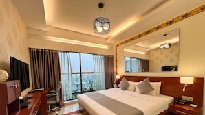 A hotel room with a large bed, a desk with a TV, a chair, a desk lamp, and a view of the city through large windows.