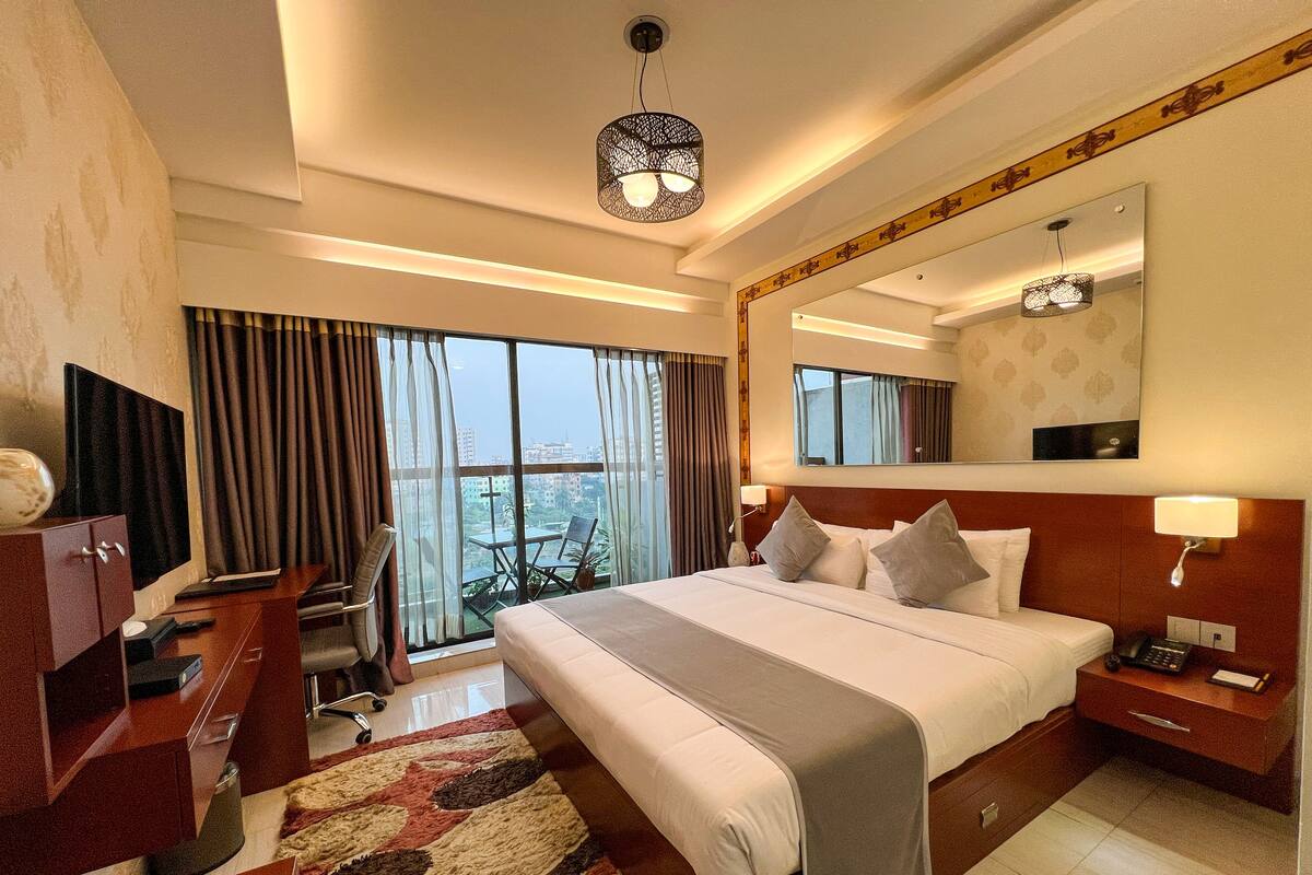 A hotel room with a large bed, a desk with a TV, a chair, a desk lamp, and a view of the city through large windows.