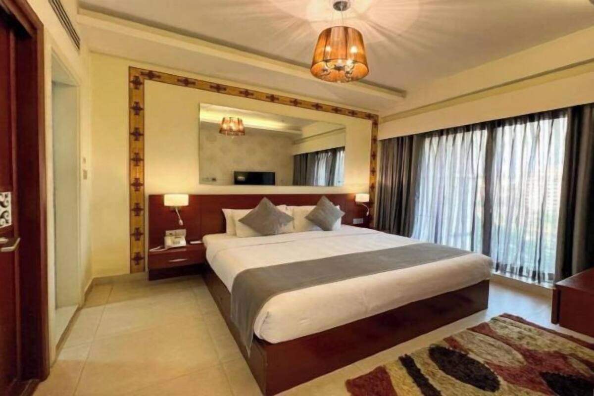A hotel room with a large bed, a bedside table, a mirror, a ceiling light fixture, and a window with curtains.